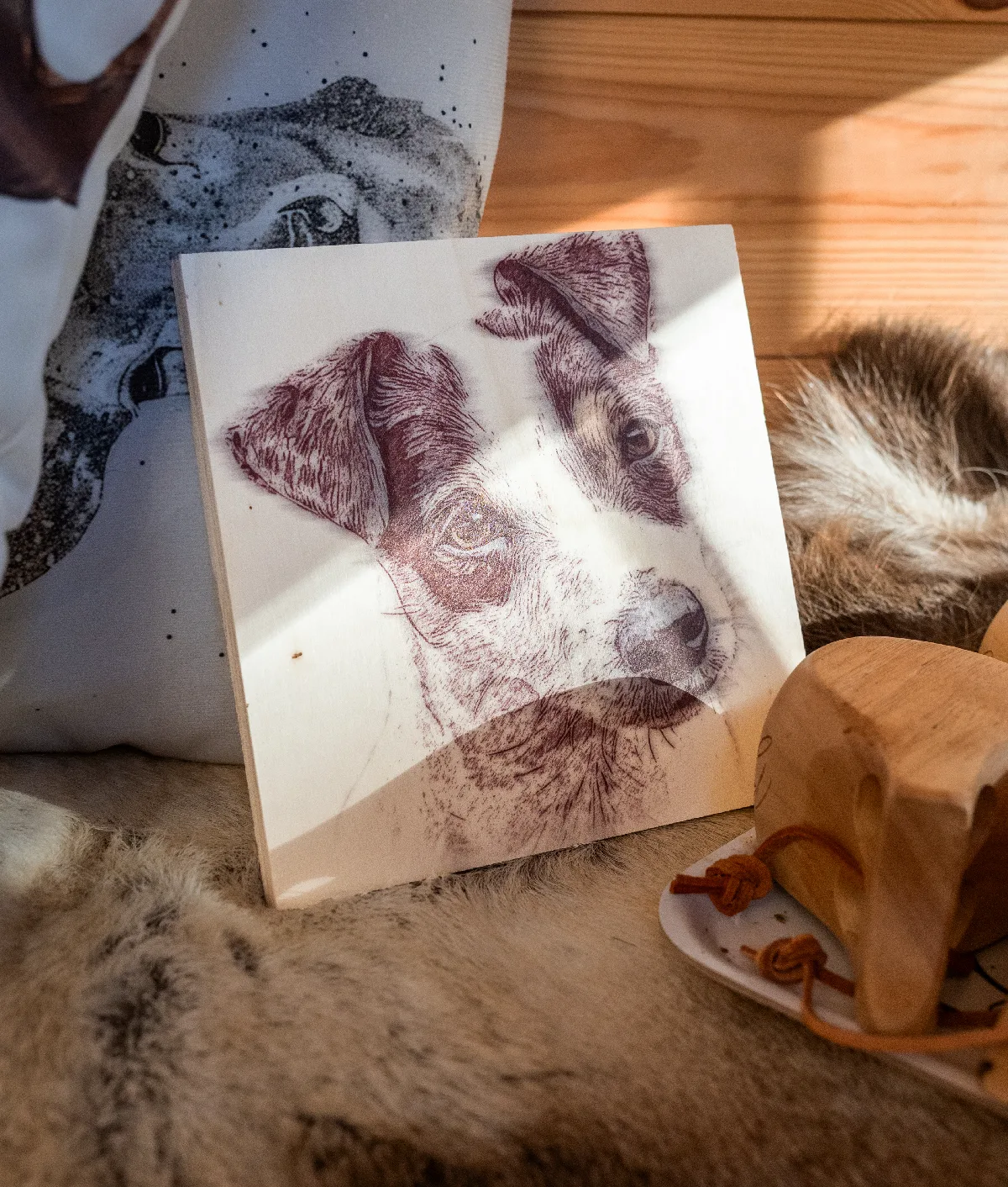 Wooden board with a wood burn style Jack Russell portrait, leaning against a dog-print cushion on a fur rug in the sunlight, next to a wooden cup with a leather cord.